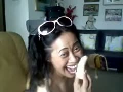 A plump Thai woman is eating a banana while flaunting her zeppelins.